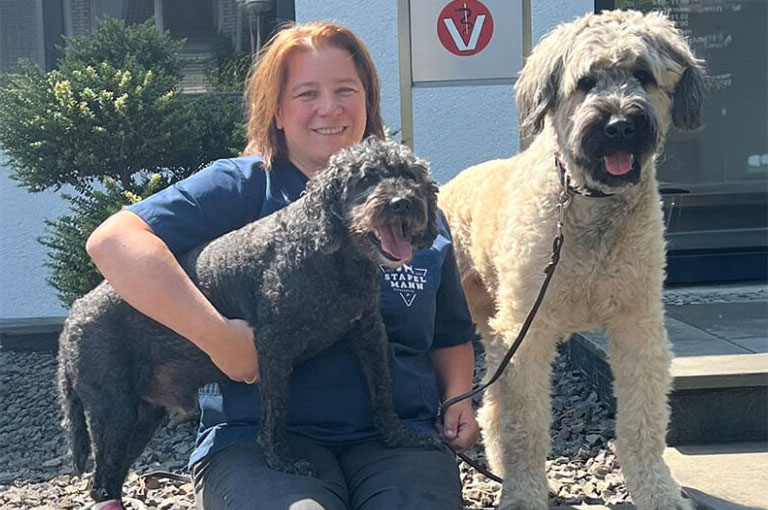 Eine lächelnde Frau hält zwei Hunde, einen schwarzen und einen beigen, vor einem Gebäude.